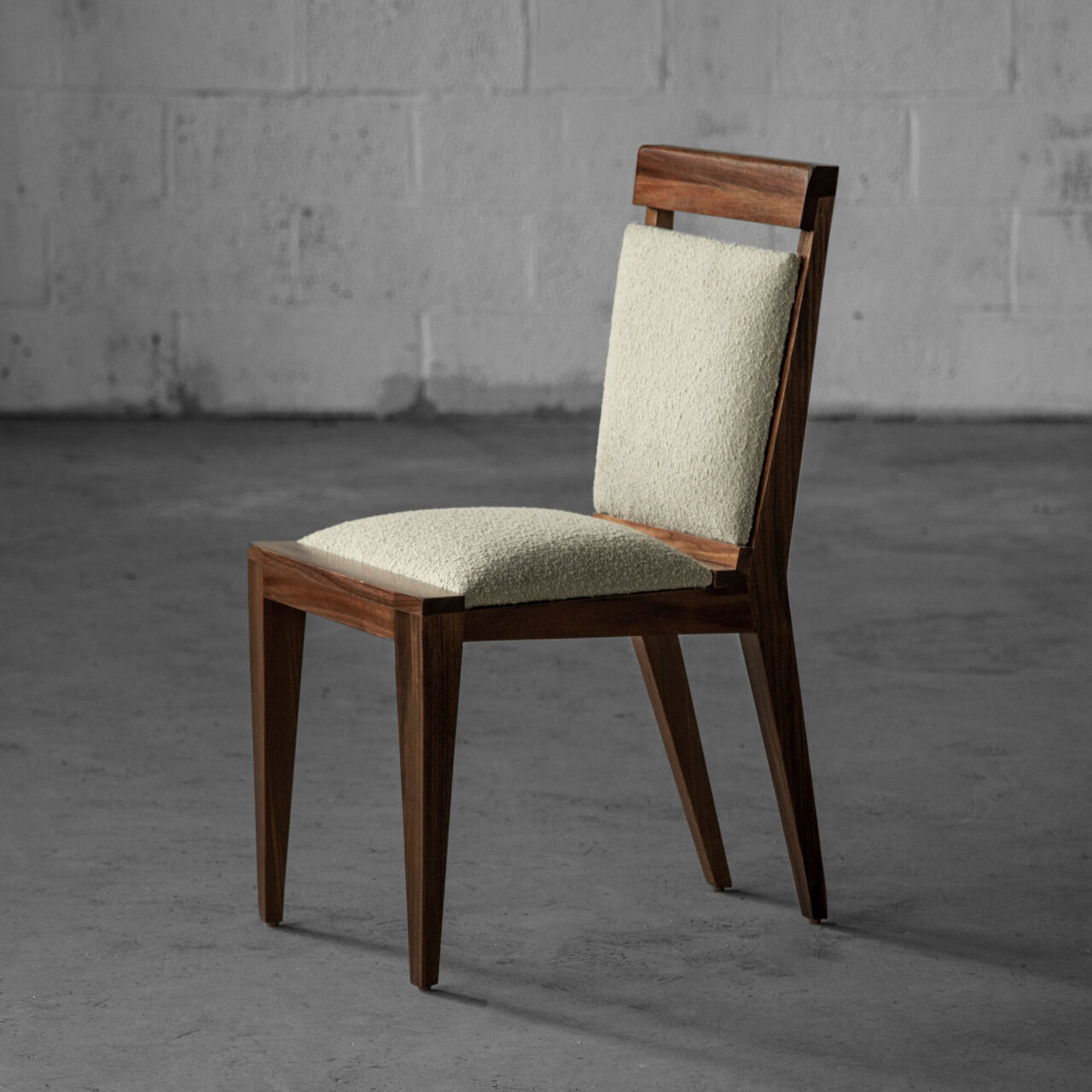 A minimalist wooden chair called Angles Chair with a light speckled cushion on the seat and backrest, set against a bare concrete background. This piece, by SENTIENT Furniture, blends modern design with a touch of comfort.
