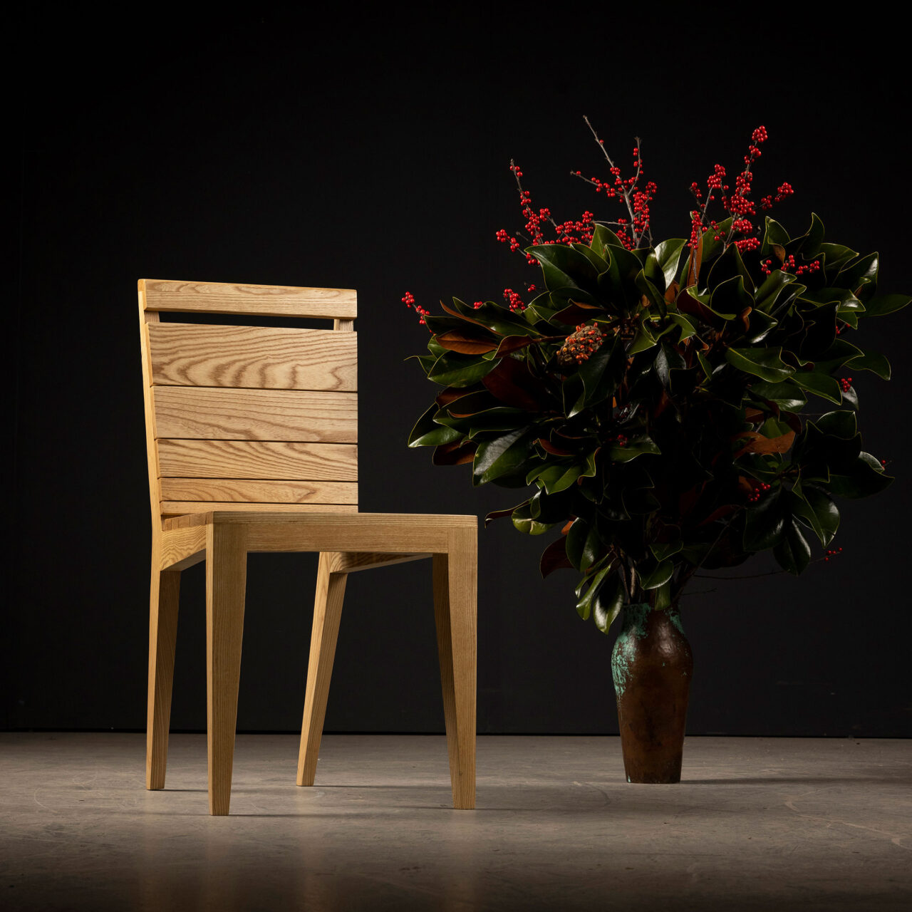 Unique dining chairs - a minimalist wooden Angels chair by SENTIENT Furniture, showcasing a natural oak finish with clear lines and a modern silhouette against a black backdrop with a large, leafy green plant and red berries to the side.