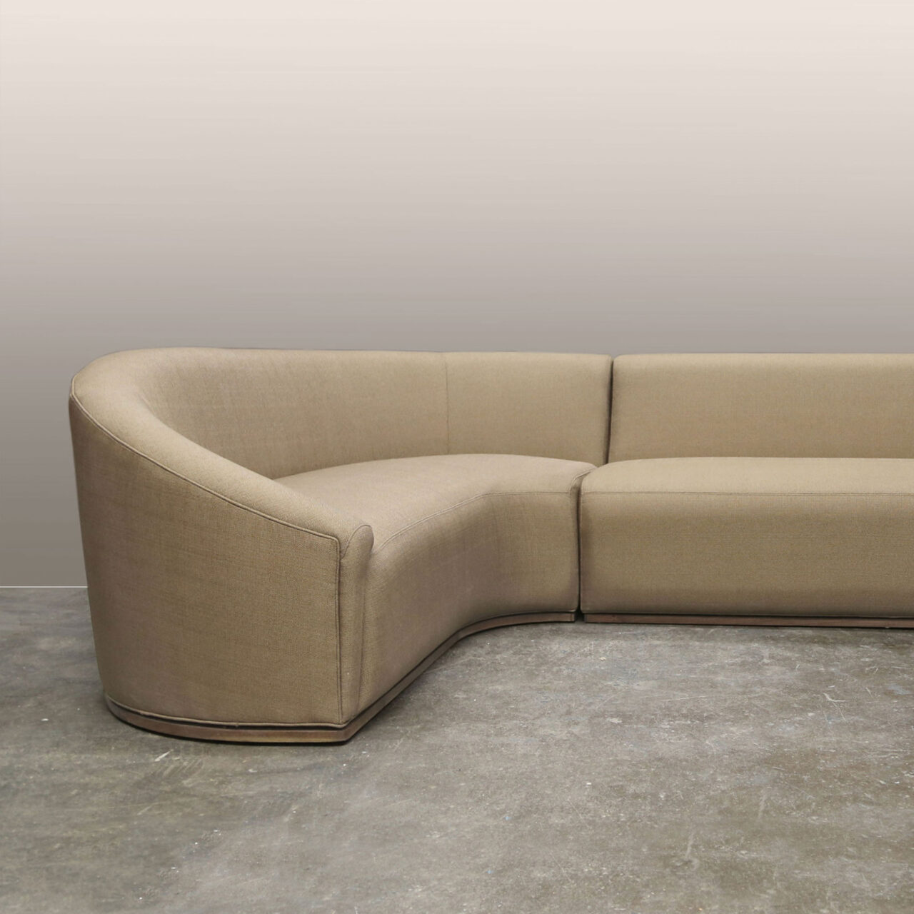 A curvilinear sectional sofa, Baashe, from SENTIENT Furniture with a beige upholstery, designed to provide a seamless and cozy seating area that contours to a room's corner, set against a simple backdrop.