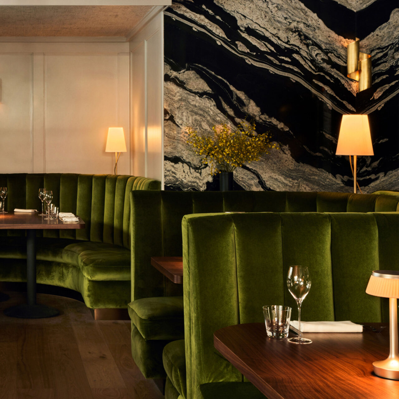 An intimate restaurant interior featuring luxurious green velvet banquette seating by SENTIENT Furniture, with a dark marbled wall accent and warm ambient lighting, creating a cozy dining atmosphere.