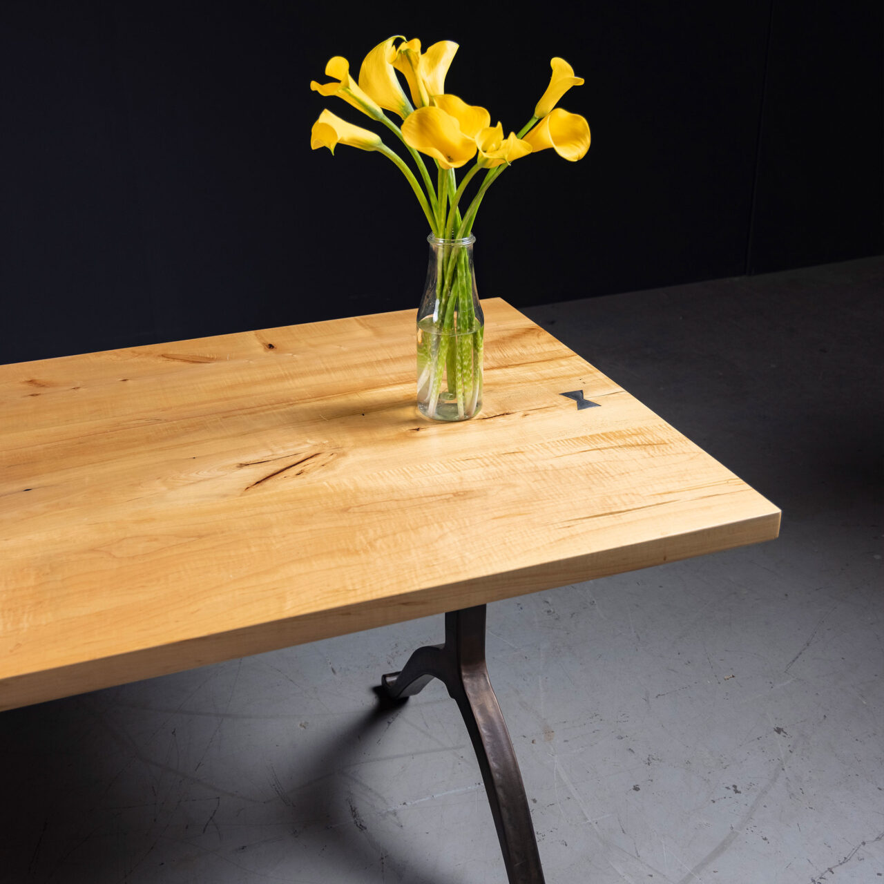 SENTIENT Furniture presents a light wood, maple, communal table featuring a minimalist design. A clear glass vase with bright yellow flowers adds a pop of color to the scene, complemented by the industrial-style metal table leg.
