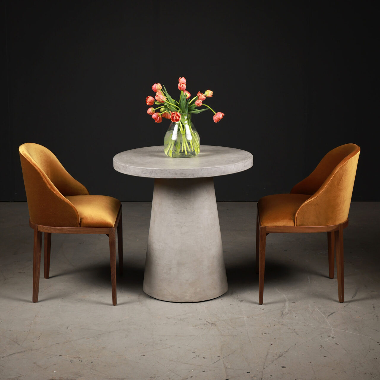 A contemporary dining setup by SENTIENT Furniture with a round concrete table and two mustard velvet chairs, complemented by a bouquet of red tulips on a dark studio background, showing the portfolio of restaurant tables by SENTIENT.
