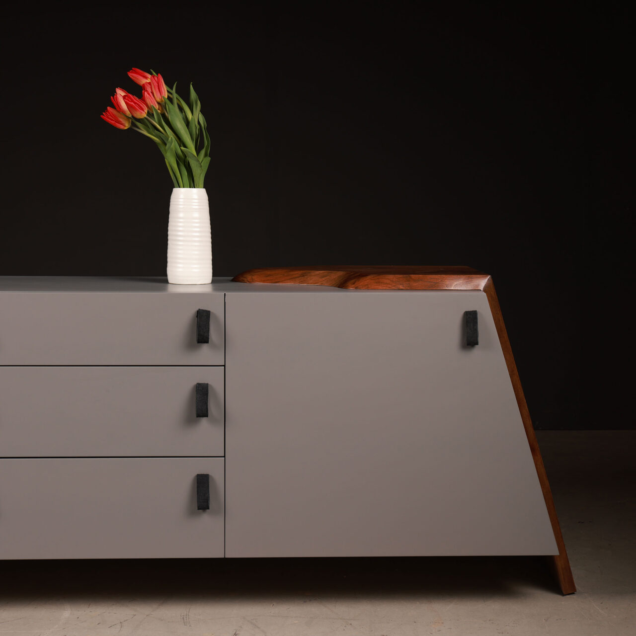 A sleek gray Luxor credenza by SENTIENT Furniture with black handles and a warm wooden top, decorated with a vase of red tulips on a dark gray surface, showcasing a blend of modern and rustic styles in luxury credenzas.