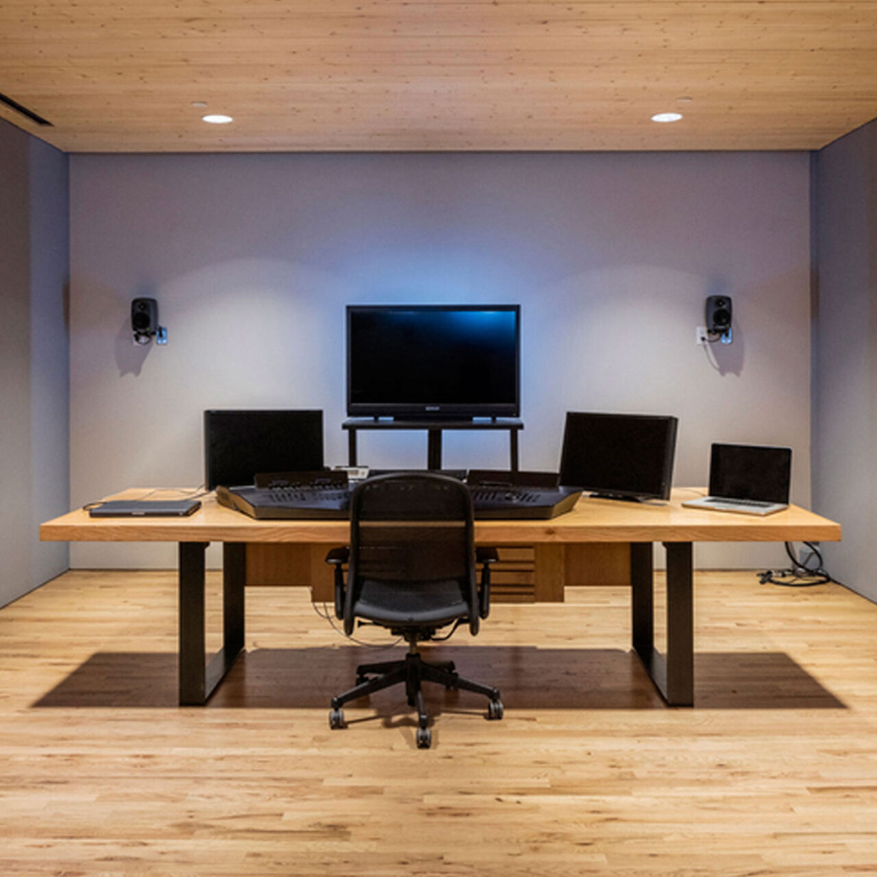 An office setup by SENTIENT Furniture featuring a custom wooden desk with ample surface area for multiple computers and audio equipment. The room has a professional ambiance with a neutral color palette and wooden ceiling.
