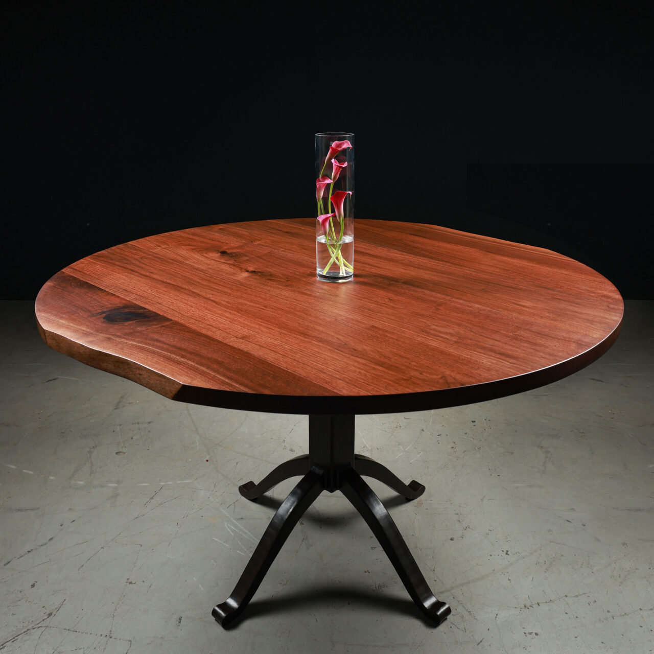 Flow, a pedestal round dining table by SENTIENT Furniture with a beautiful reddish wood top and a black metal base. The table is adorned with a tall glass vase containing red and green flowers, set against a black backdrop for a dramatic effect.