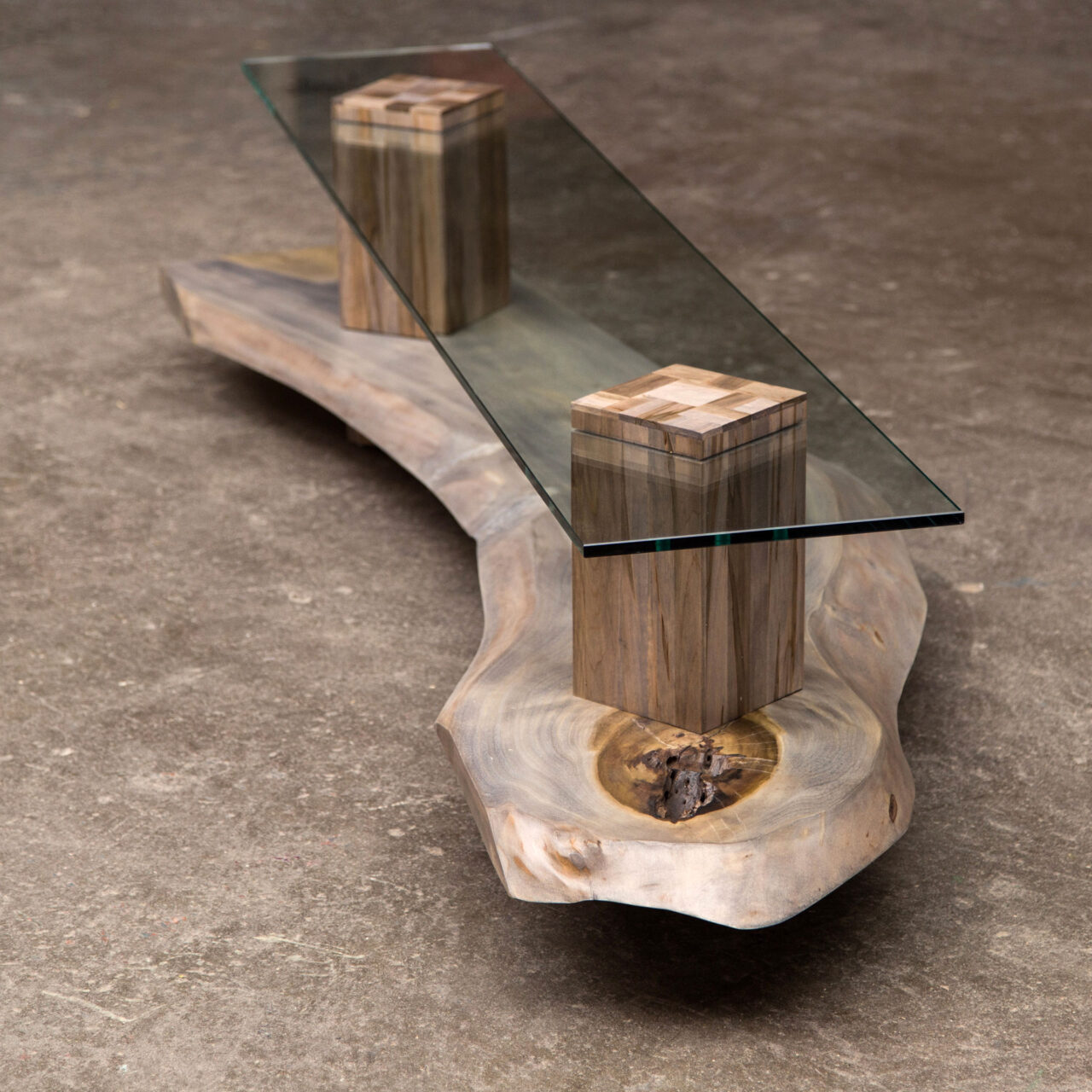 A unique luxury console table by SENTIENT, Foothills, combining a live-edge wood slab and a marble slab with a clear glass top, placed on geometric walnut pedestals on a sidewalk.