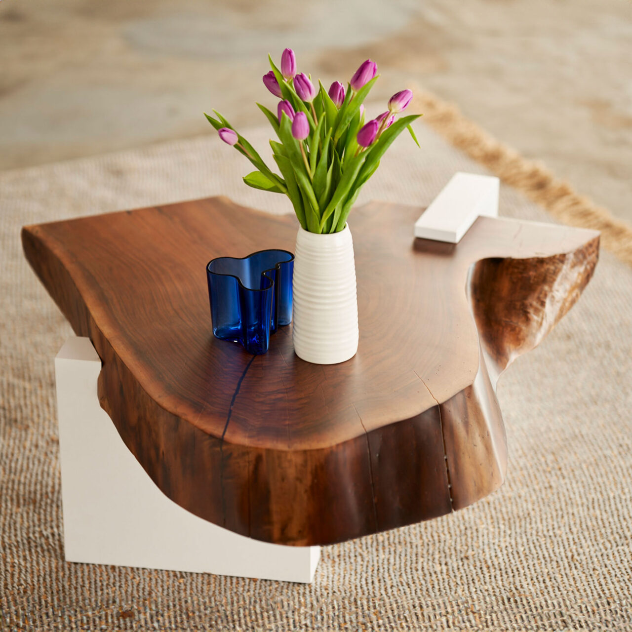 Elegant SENTIENT Furniture live-edge coffee table crafted from a single walnut slab, featuring natural contours and a minimalist white base, adorned with a vase of purple tulips.