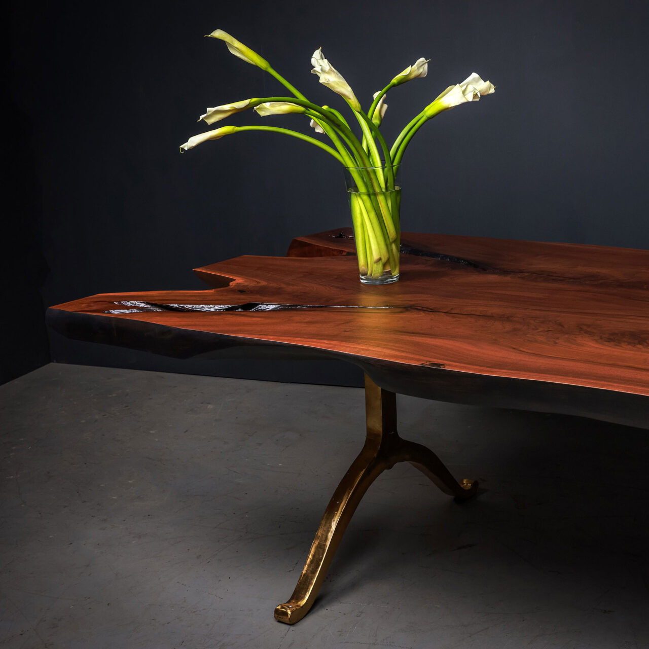 A luxury live edge table by SENTIENT, with stunning wishbone legs, and organic, raw tabletop, with a vase of lilies in front of a black background in a lit studio.