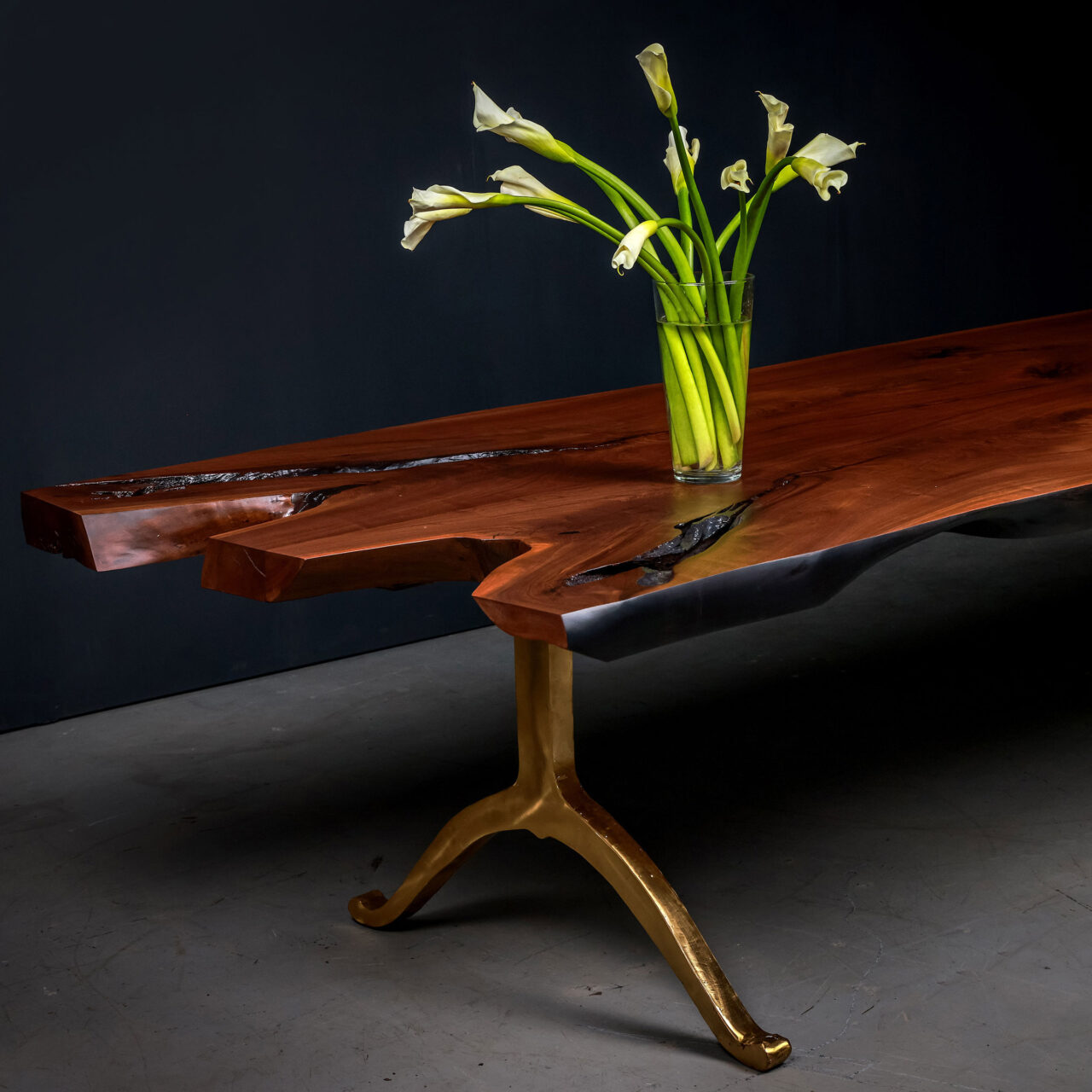 A luxury live edge dining table by SENTIENT Furniture, with a rich wooden top and contrasting golden metal legs, elegantly adorned with a glass vase of white calla lilies.