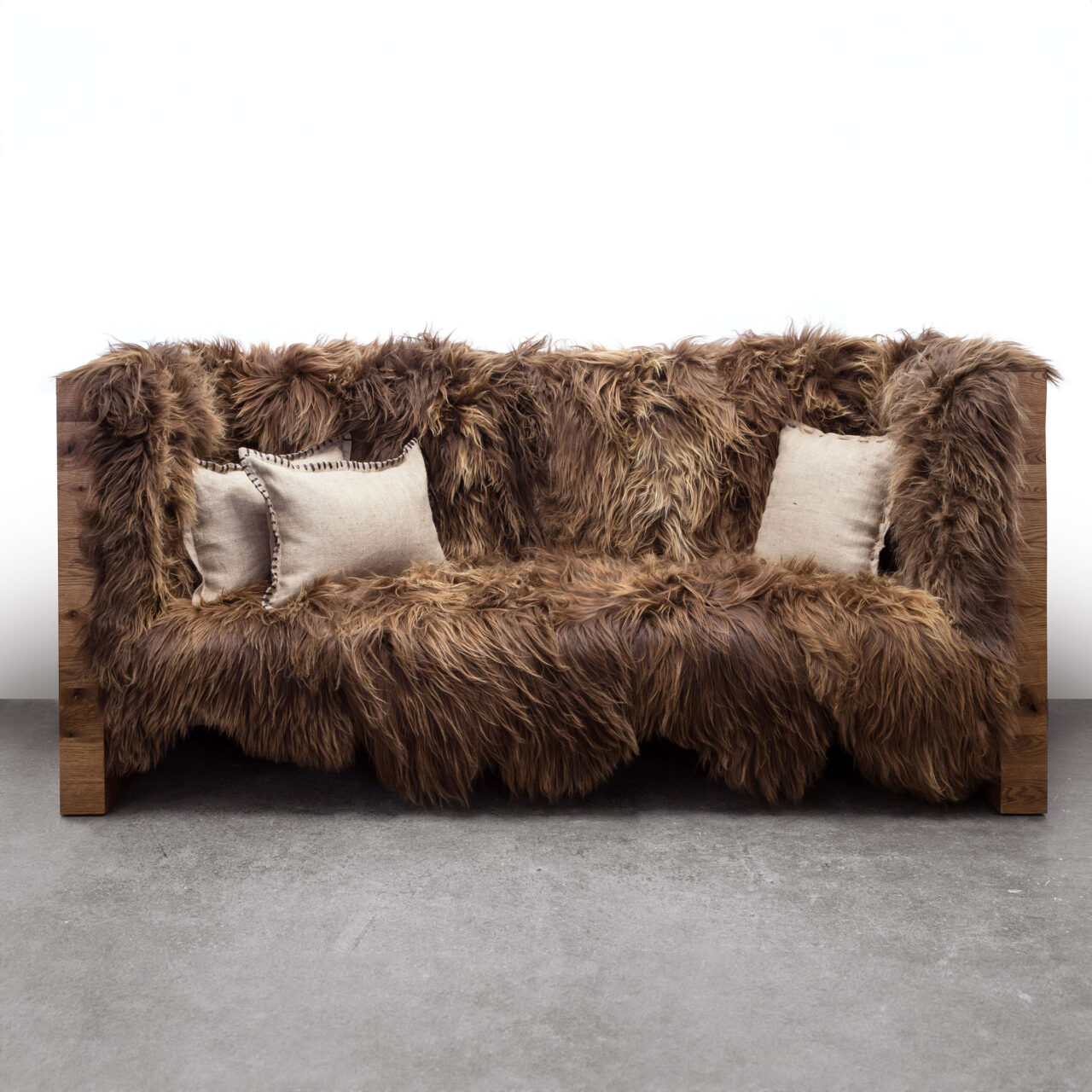 A luxurious SENTIENT long wool sofa with a dense, shaggy brown fur cover and two off-white accent pillows against a plain background.