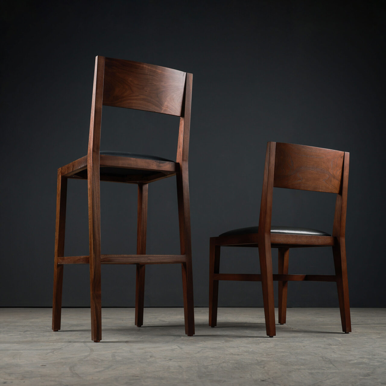 A pair of high-backed luxury solid wood unique dining chairs by SENTIENT Furniture, crafted with rich walnut wood and black leather upholstery, presented in a dramatic studio setting with a black background.