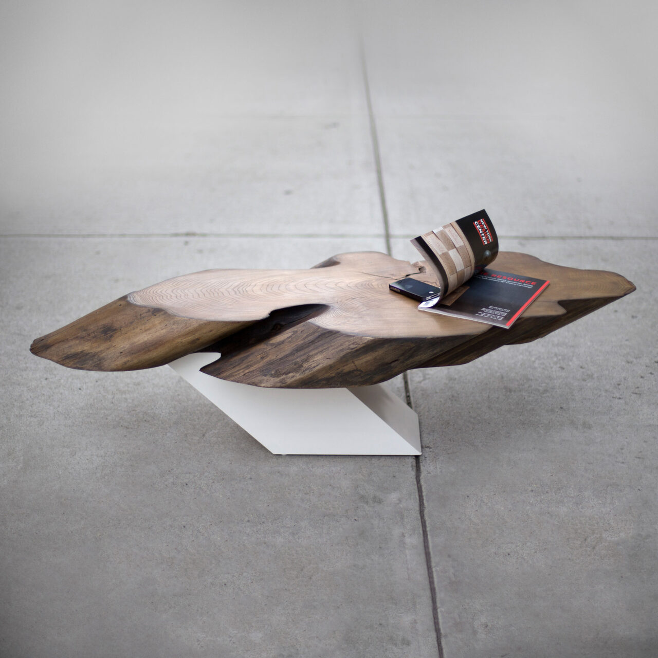 SENTIENT Furniture's 'Zora Coffee Table', an organic-shaped wooden tabletop on a minimalist white stand, with books and a phone on top, in a modern concrete space.